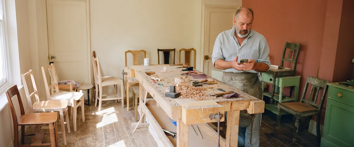 Photo d'illustration : un artisan restaurateur de meubles regarde son téléphone dans son atelier, l'air soucieux.