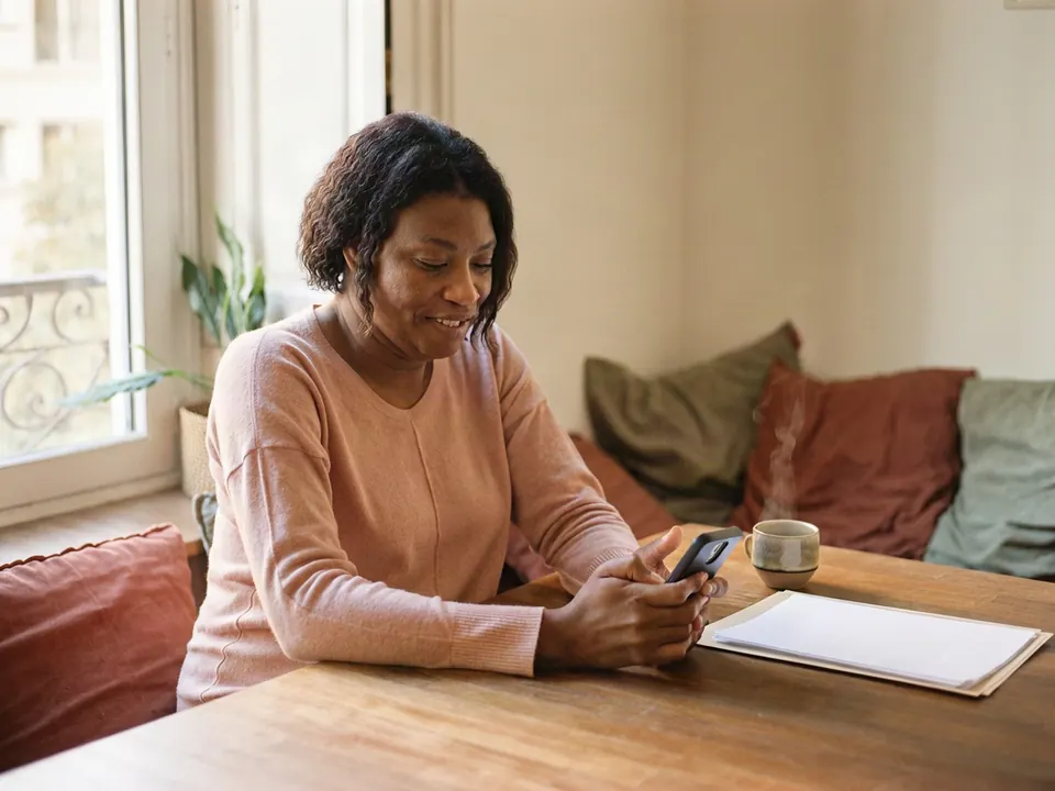 Photo d'illustration : une femme lit un message sur son téléphone et sourit, soulagée, une tasse de thé à côté d'elle.